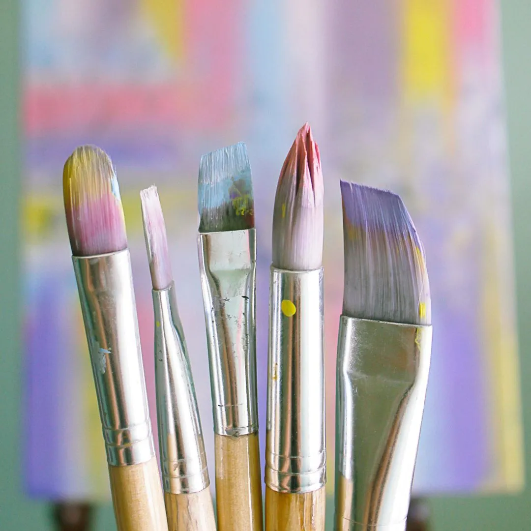 Five paint brushes arranged in front of a colorful painting on canvas, showcasing various brush sizes and styles.