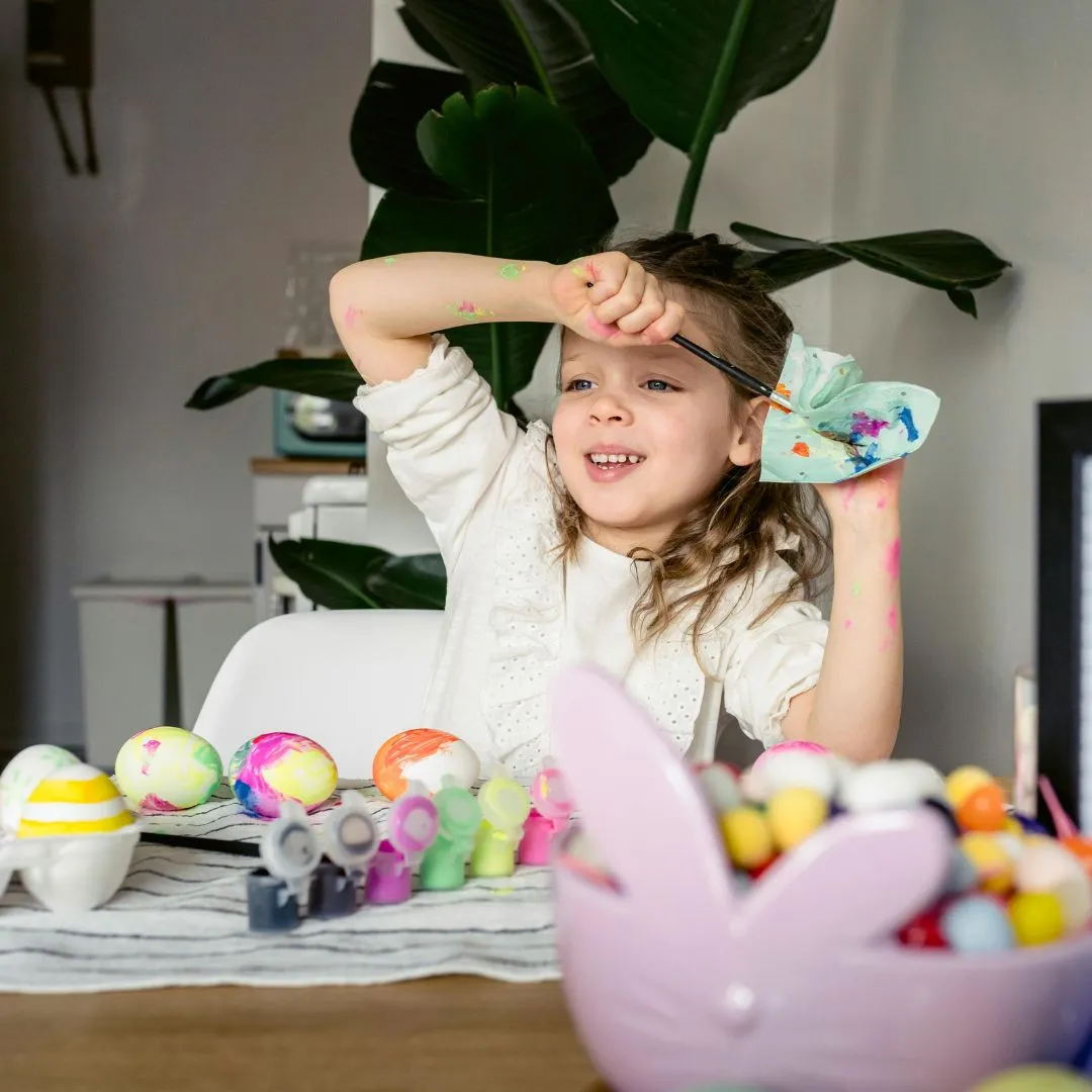 A little girl smiles while holding an Easter egg in front of a table filled with colorful Easter eggs.