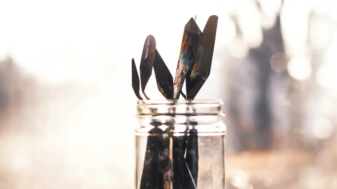 A jar filled with various paintbrush palette knives, their colorful, worn edges reflecting light against a blurred background.