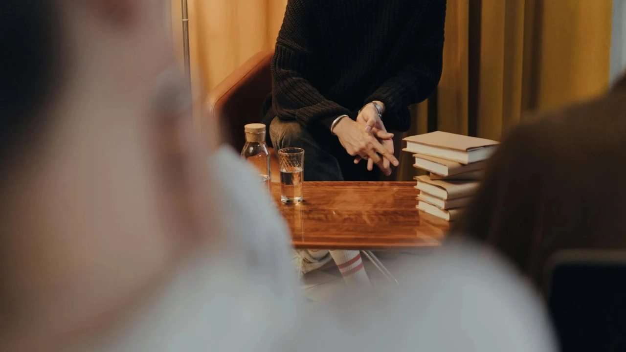 A person in a black sweater sits on a chair, hands clasped on a wooden table with books and a glass of water beside them.