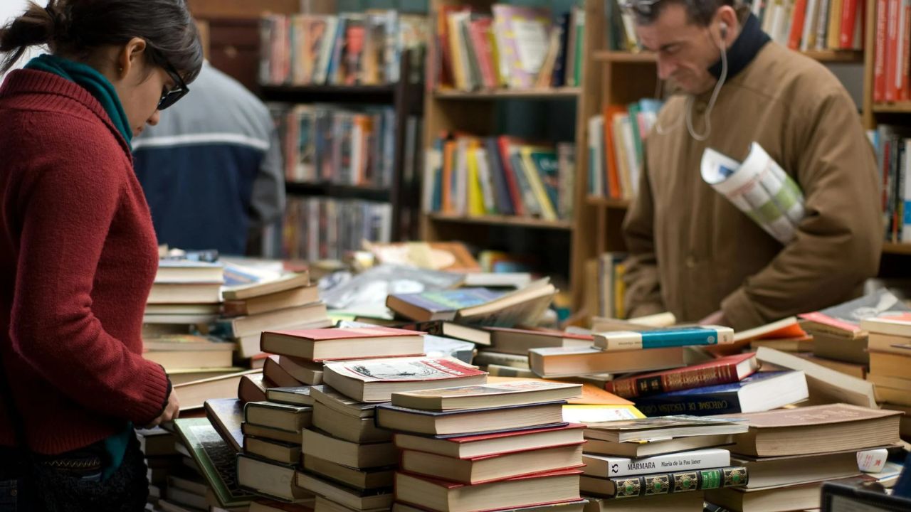 A woman in a red sweater and a man browse a cluttered table filled with books in a cozy bookstore. Bookshelves line the background.