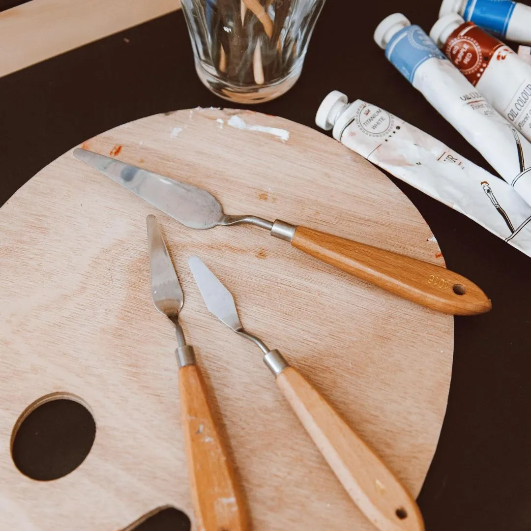 Three artist's palette knives with wooden handles rest on a circular wooden palette, surrounded by paint tubes and a glass of brushes.