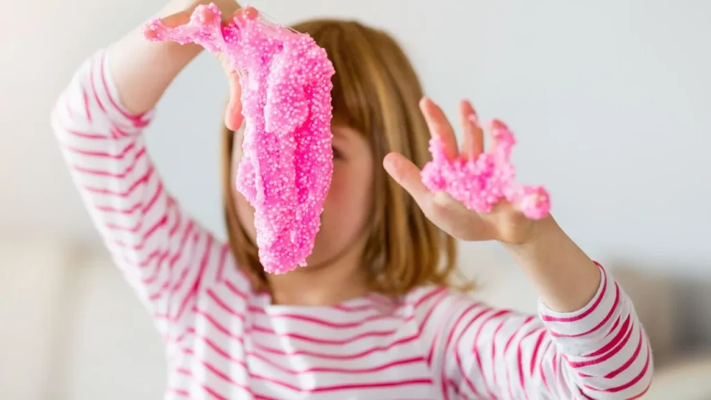 A young girl joyfully plays with bright pink slime, stretching and squishing it in her hands.