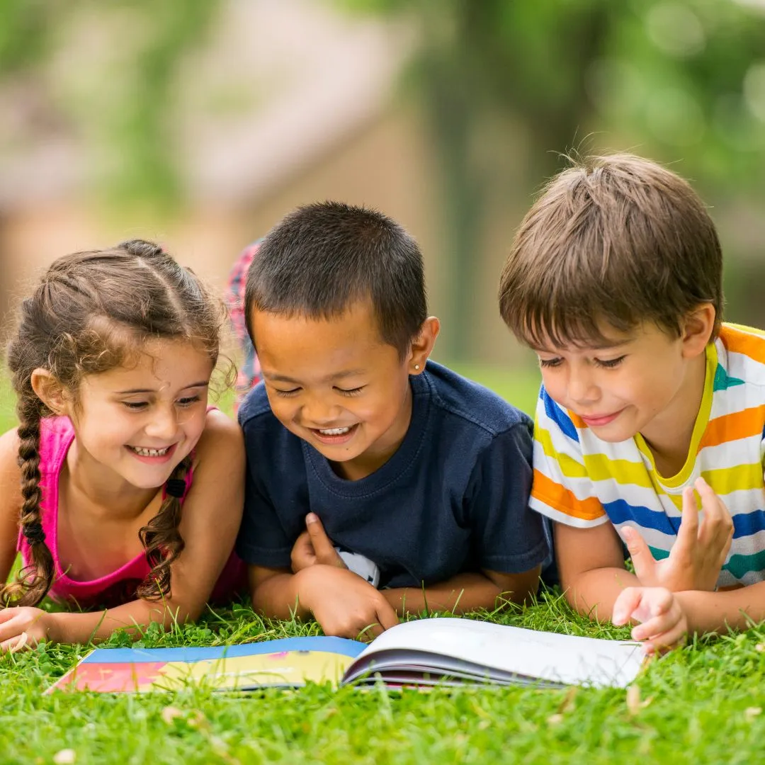 Three children sitting on the grass, engrossed in reading a book together, surrounded by a sunny outdoor setting.