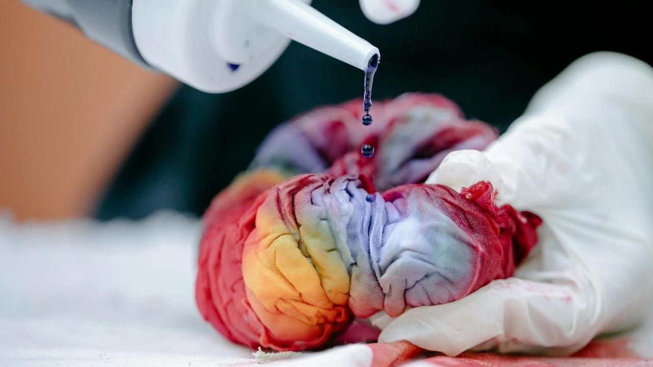 A person wearing a white glove pours dye onto a piece of fabric, creating vibrant colors for a textile project.