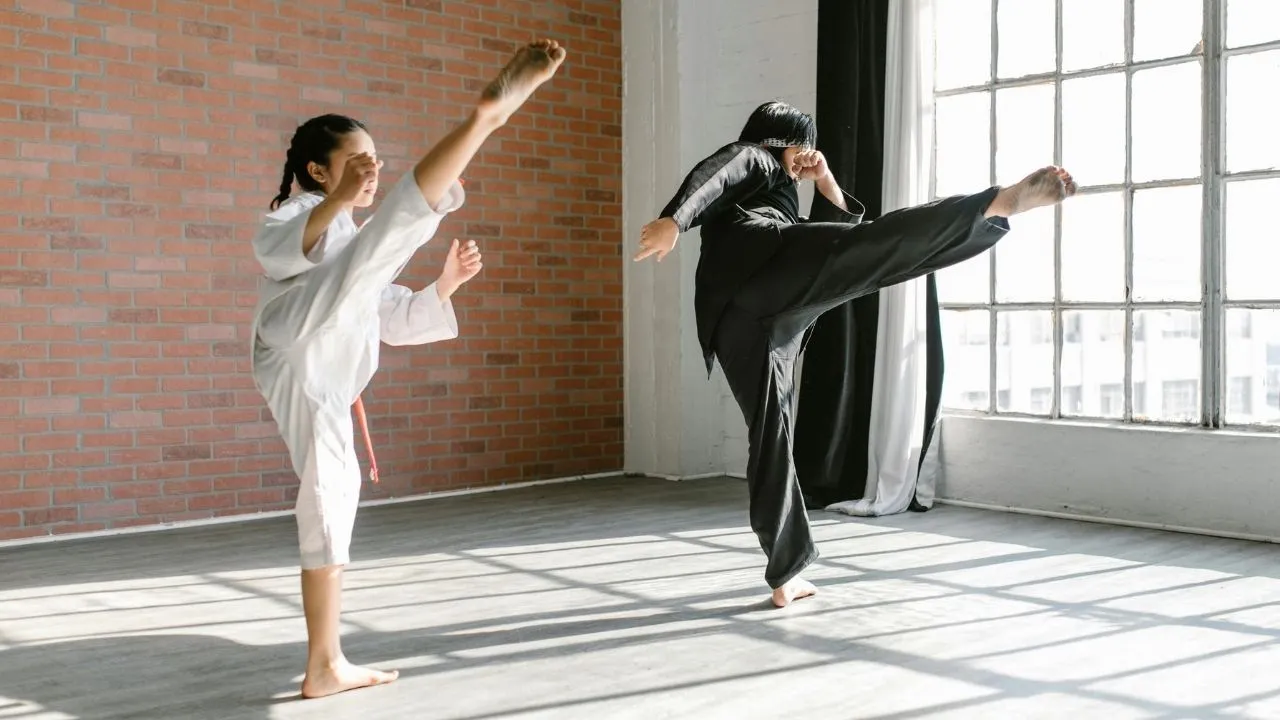 Two individuals in jiu jitsu uniforms practicing techniques in a spacious, empty room.