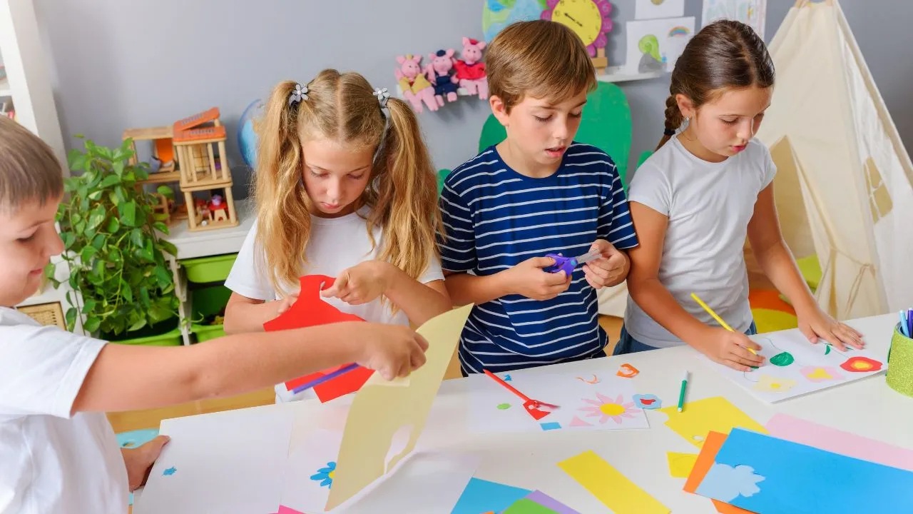 Children engaged in making paper crafts at desks in a bright classroom filled with art supplies and colorful decorations.
