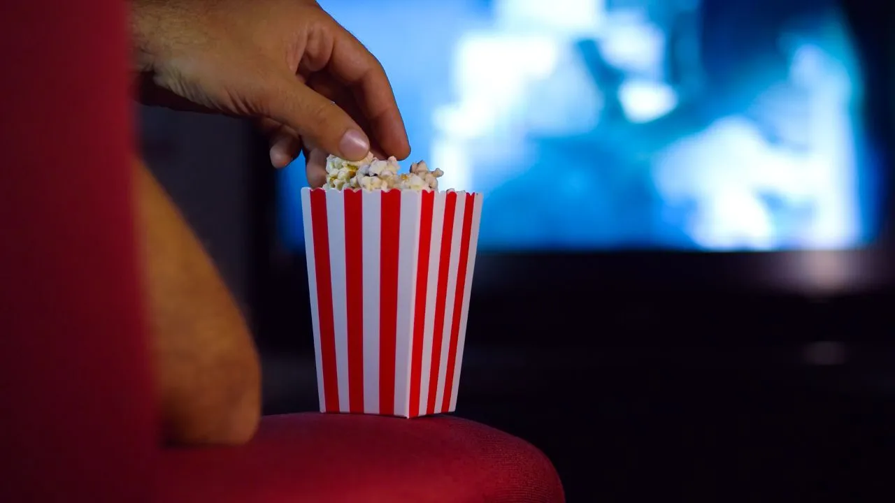 Hand reaching into a red and white striped popcorn box, set against a blurred movie screen background. Cozy, cinematic atmosphere.