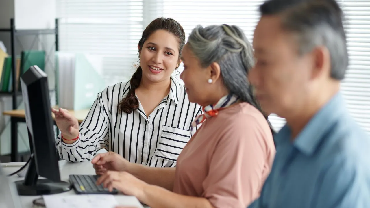 A young woman in a striped shirt explains something on a computer to an older woman in a pink blouse. An older man in blue is in the foreground, blurred. The mood is collaborative and supportive.