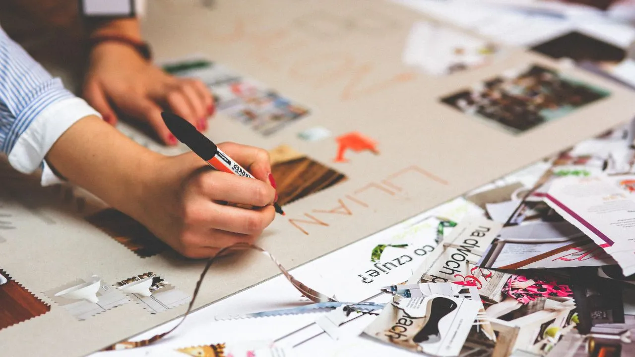 A hand writes with a black marker on a large paper surrounded by colorful magazine cutouts and swatches, suggesting a creative, brainstorming session.