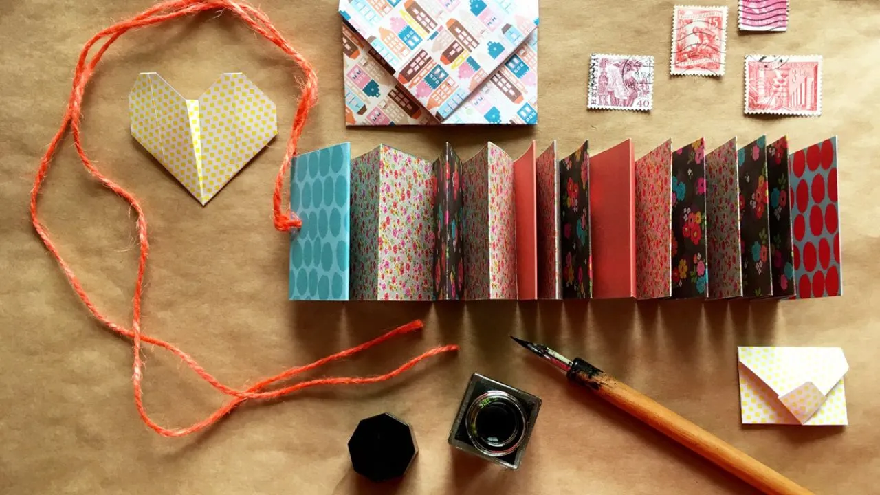 Crafting scene with colorful origami, mini envelope, stamps, an accordion-style scrapbook, red string, inkwell, pen, on brown paper. Creative, playful feel.