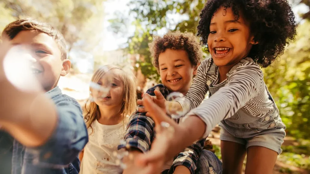 Four joyful children reaching towards the camera, playing outside on a sunny day. They are surrounded by greenery, with expressions of excitement.