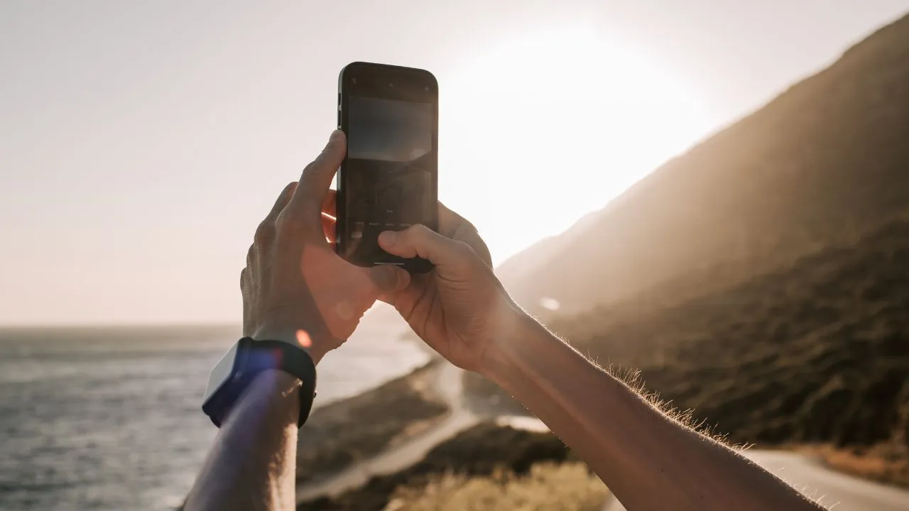 A person holds a smartphone up towards a bright sunlit sky, capturing a scenic coastal view. The scene conveys a sense of travel and adventure.