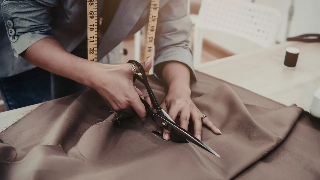 A tailor with a measuring tape around their neck is cutting brown fabric with scissors on a table. The scene conveys focus and craftsmanship.