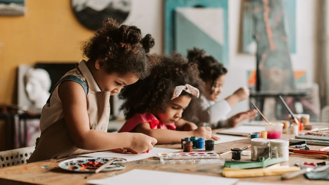 Three young children are painting at a wooden table, focused and engaged. Art supplies are scattered around, creating a lively and creative atmosphere.