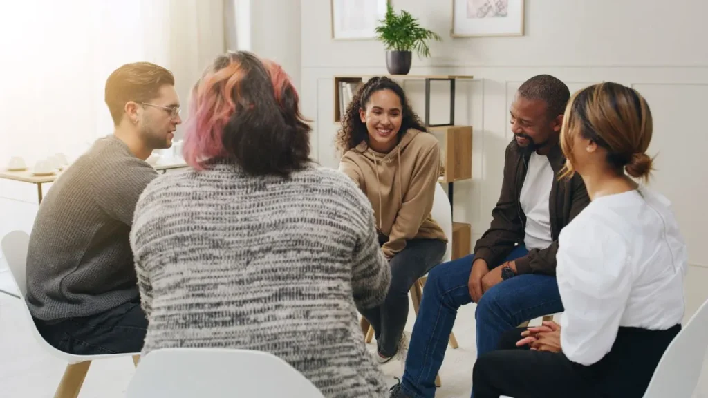 A diverse group of six people sit in a circle during a casual meeting in a bright room, smiling and engaged, conveying warmth and friendliness.