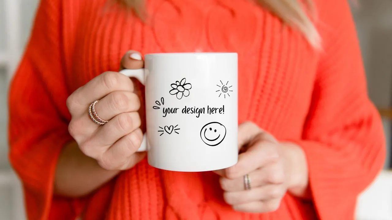 A person wearing an orange sweater holds a white mug with doodles and the text "your design here!" printed on it.