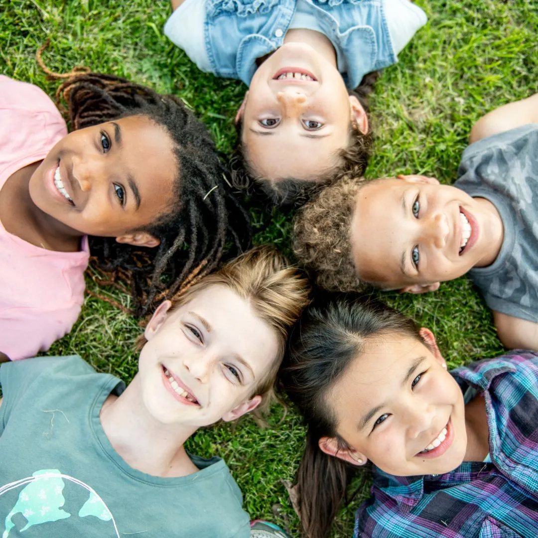 Five diverse children lie on grass in a circle, heads together, smiling joyfully at the camera. The scene conveys friendship and happiness.