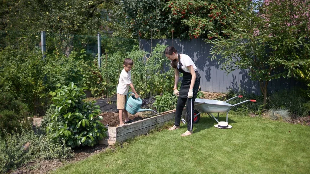 A woman and a child garden together on a sunny day. The child waters a raised bed, while the woman stands nearby with a wheelbarrow, surrounded by greenery.