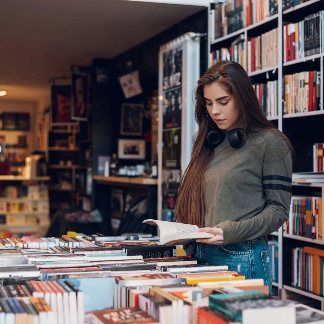 Young woman with headphones around her neck reads intently in a cozy bookstore. Bookshelves filled with colorful books create a warm atmosphere.