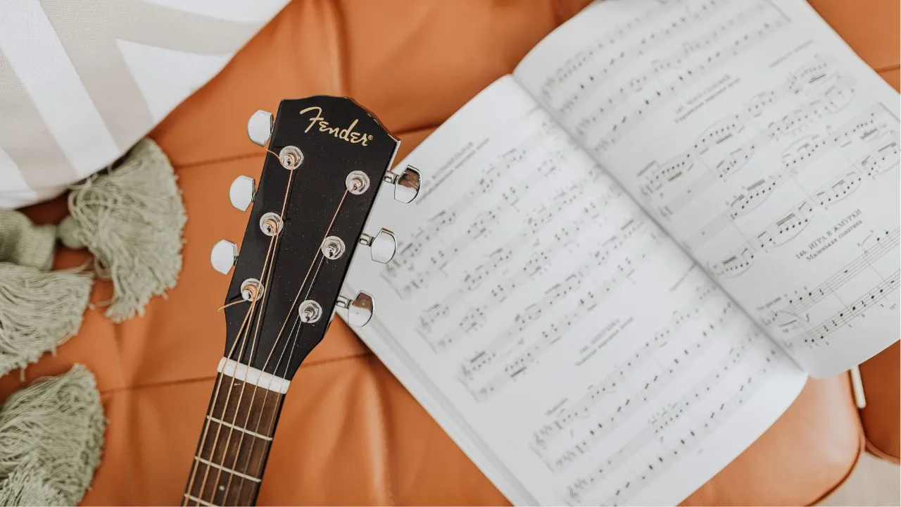 Close-up of a Fender guitar headstock resting on an orange sofa beside an open sheet music book, conveying a relaxed musical vibe.
