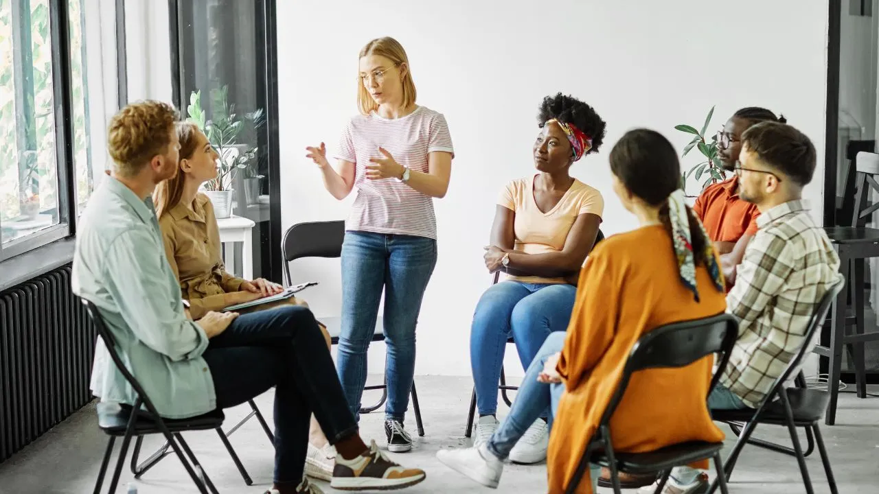 A group therapy session with six diverse individuals seated in a circle, attentively listening to a blonde woman standing and speaking, conveying a supportive atmosphere.