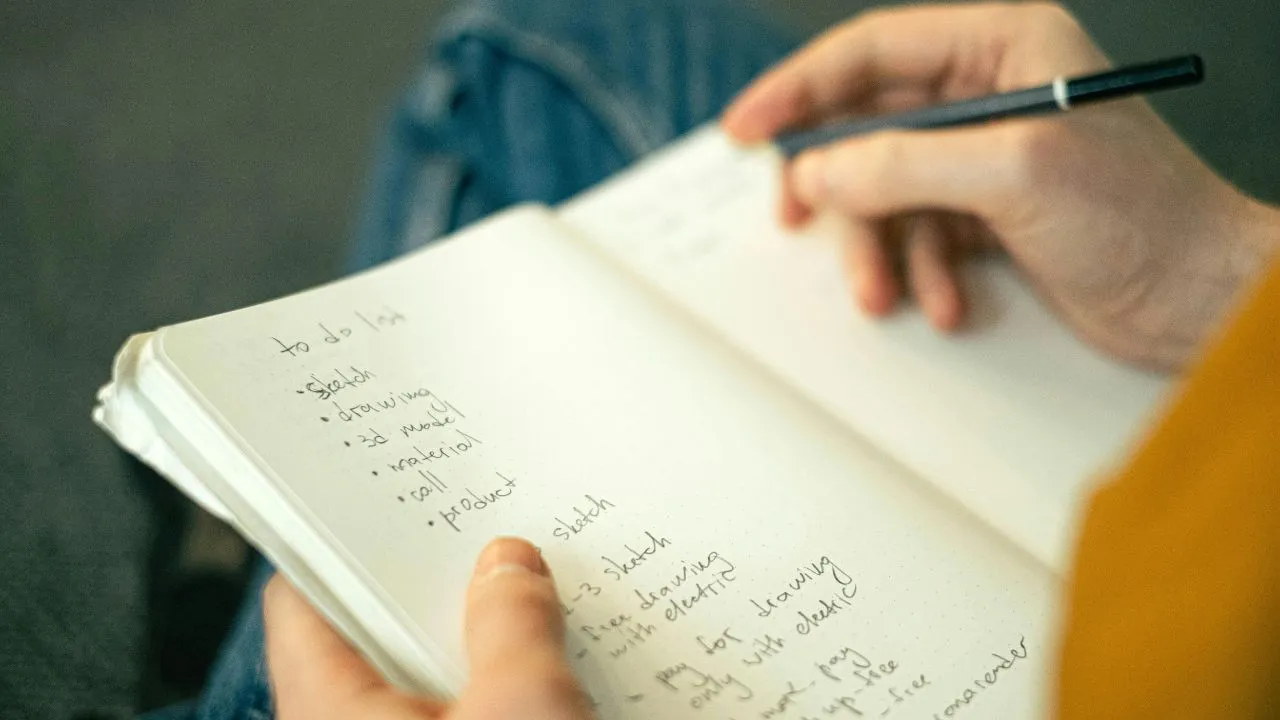 A person writes in a notebook titled "to do list" with tasks like sketch, drawing, and reading. The casual setting conveys focus and organization.