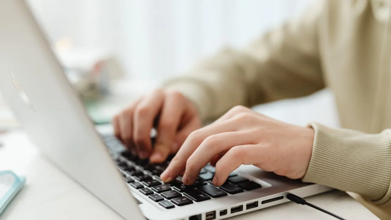 Close-up of hands typing on a laptop with a silver finish, conveying focus and productivity. Person wears a beige sweater, creating a warm tone.