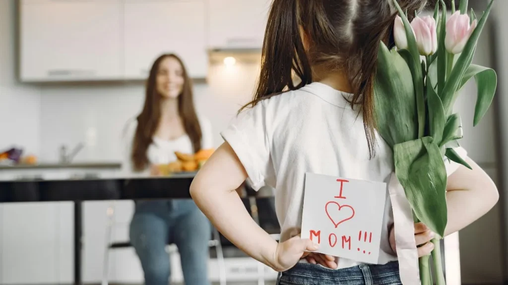 A young girl hides pink tulips and a handmade "I ❤️ Mom!!!" card behind her back, facing her smiling mother in a modern kitchen, conveying love and surprise.