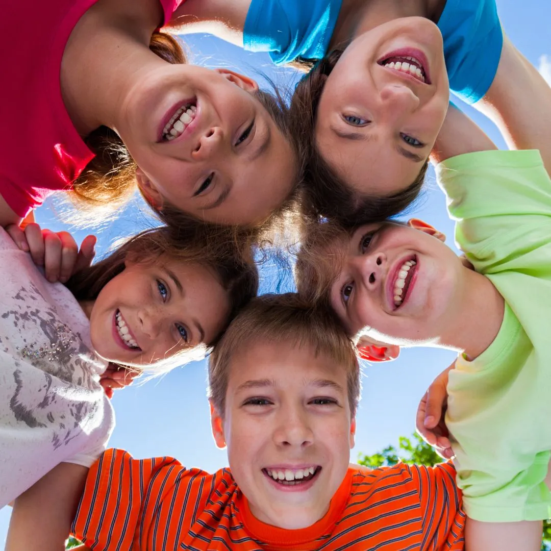 Five children form a circle, heads together, smiling joyfully at the camera. Bright clothes and a clear blue sky create a vibrant, cheerful scene.