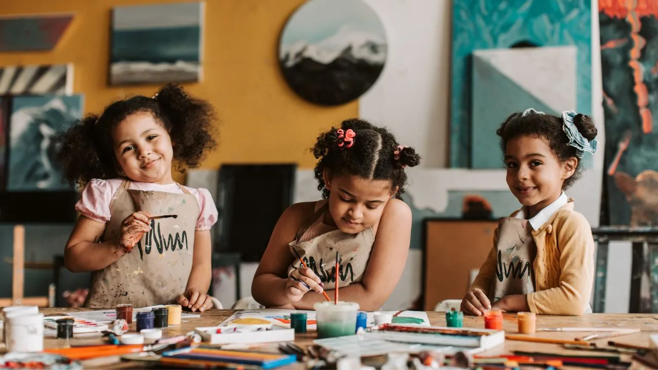 Three children sit at a table, engaged in painting with various art supplies scattered around them.