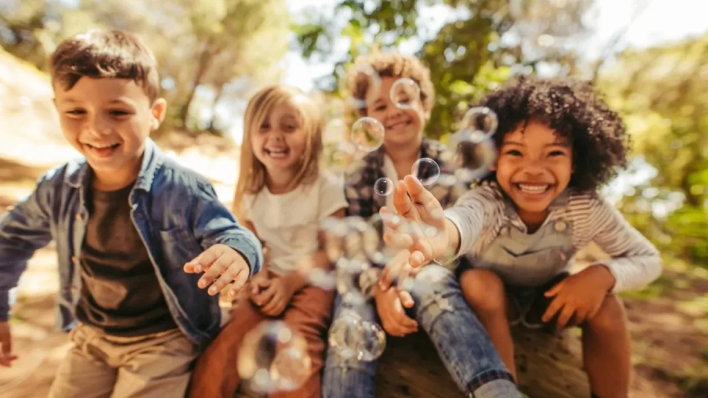 Four children sit outdoors on a sunny day, smiling and reaching for floating bubbles. Trees and sunlight create a joyful and playful atmosphere.
