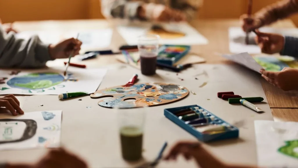 Children's hands painting at a table full of art supplies, including paints, brushes, and palettes. Vibrant creativity and teamwork atmosphere.