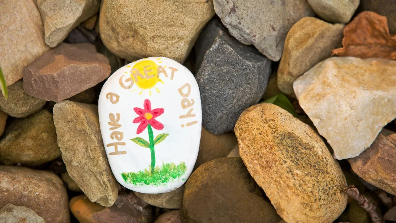 A white rock painted with a red flower and green stem says "Have a GREAT Day!" in cheerful colors. It sits among various natural stones, evoking positivity.