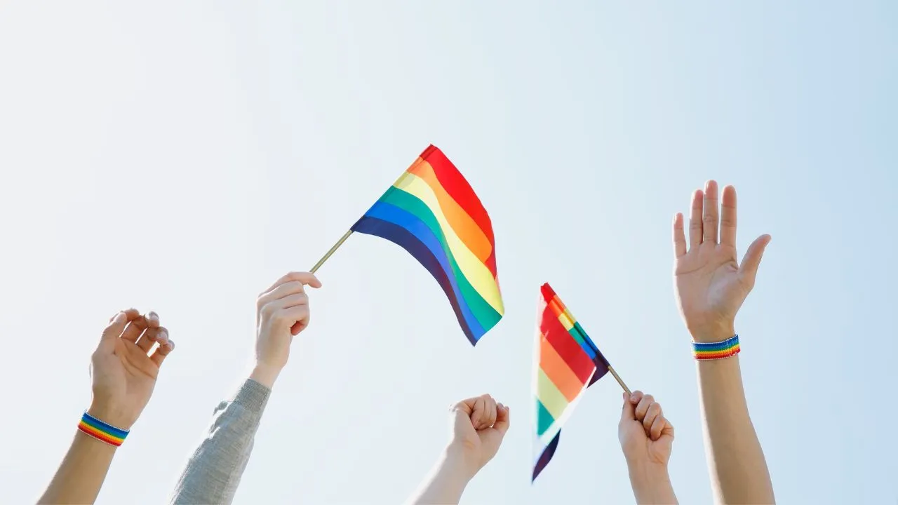 Hands of diverse individuals raise rainbow flags against a clear blue sky, wearing rainbow bracelets, symbolizing unity and LGBTQ+ pride.