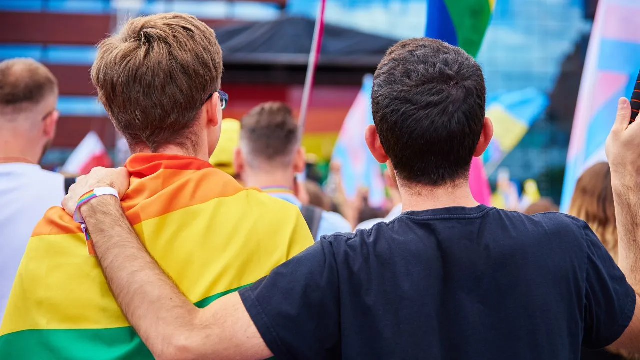 Two people with their backs to the camera embrace at a colorful outdoor event. One wears a rainbow flag, symbolizing LGBTQ+ pride, in a celebratory atmosphere.