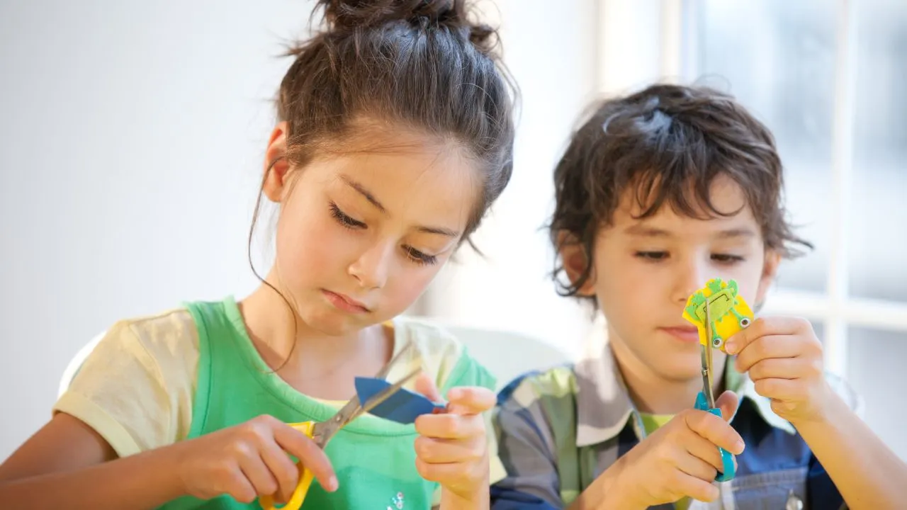 Two children are crafting paper flowers using scissors at a table, focused on their creative project.