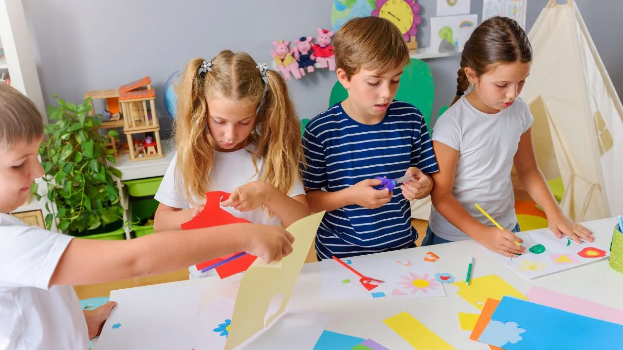 Children engaged in making colorful paper crafts at desks in a bright classroom filled with art supplies.