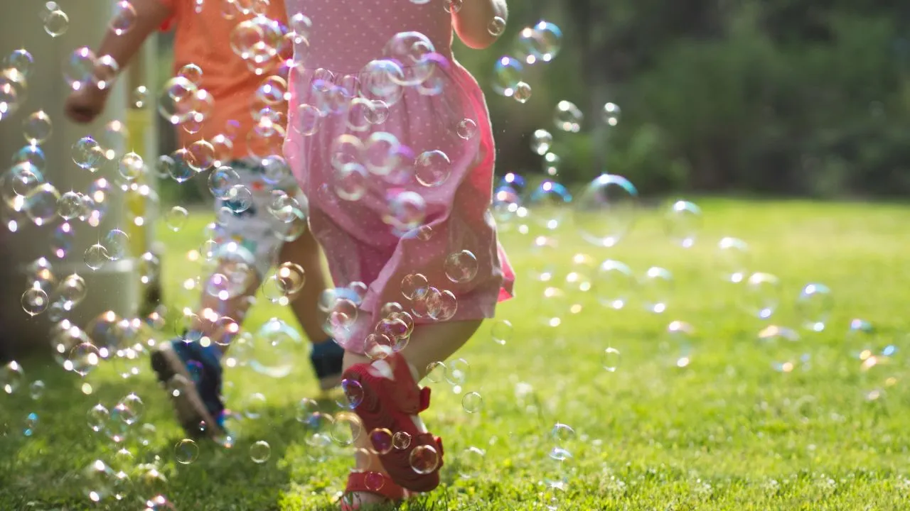 Two children joyfully run through a grassy field, surrounded by floating bubbles in the sunlight.
