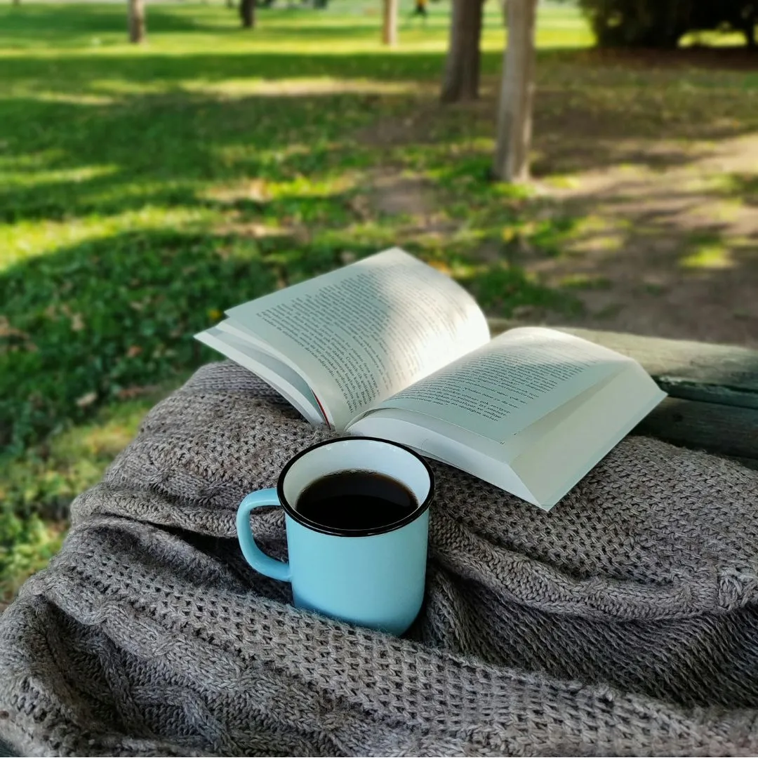 A cup of coffee and an open book resting on a blanket spread out on grass in a park setting.