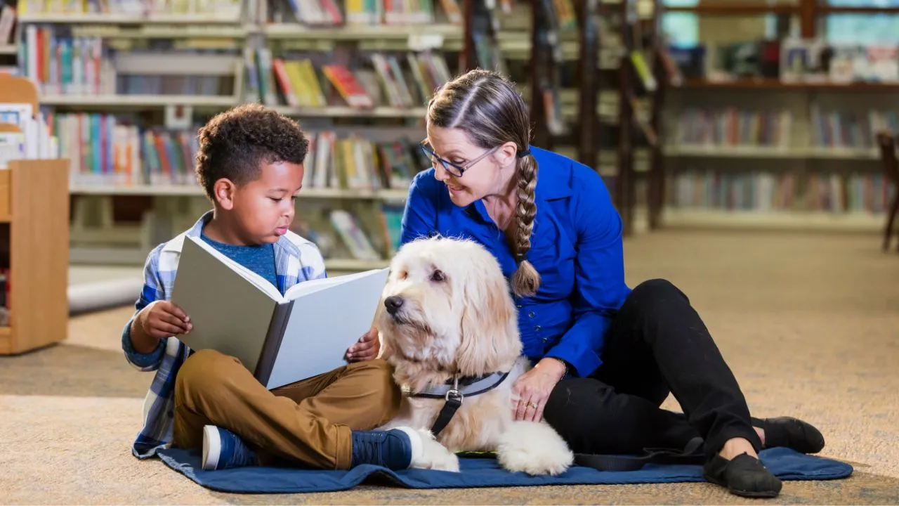 A woman and a boy read a book to a dog in a cozy library setting, surrounded by shelves of books.