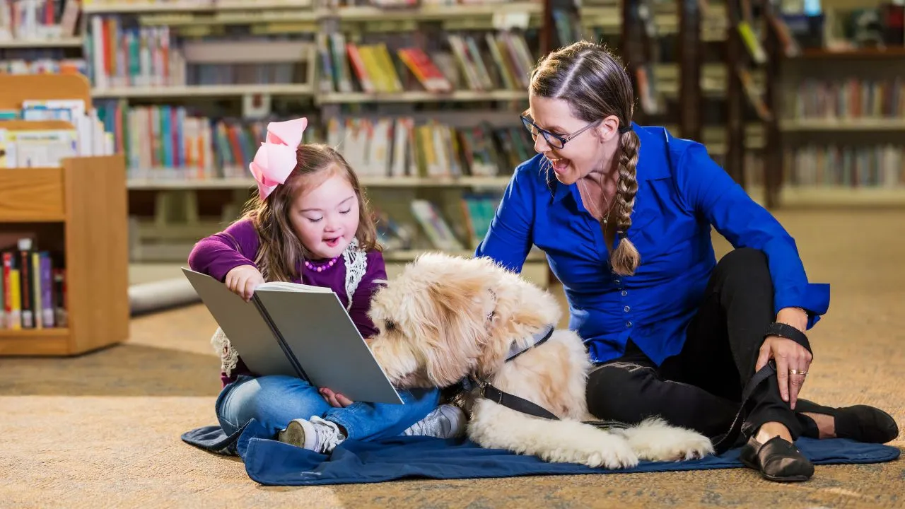 A woman and a girl sit on the floor of a library, reading a book with a dog beside them.