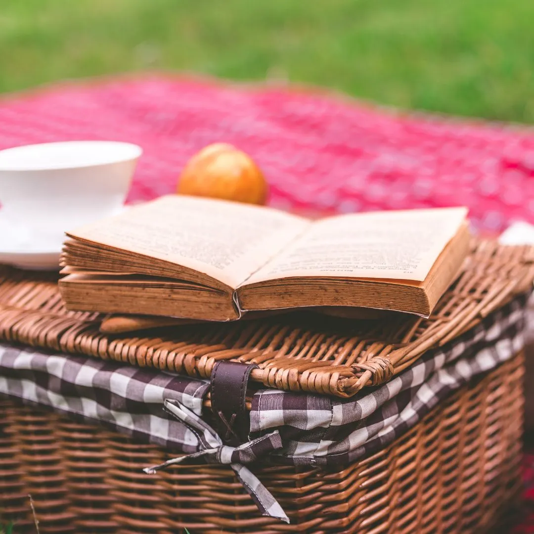  A picnic basket, a book, a cup of tea, and an orange arranged on a blanket in a park setting.