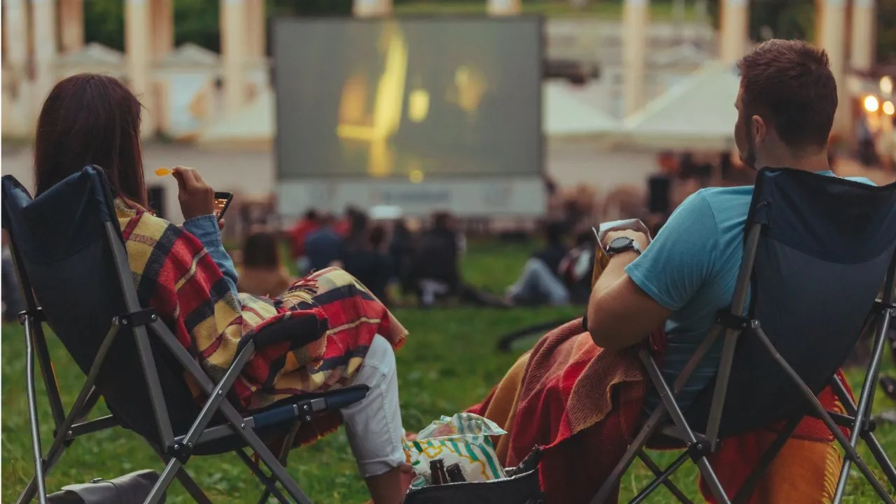 Two individuals sit in lawn chairs, watching a film on a big screen during an outdoor movie night.