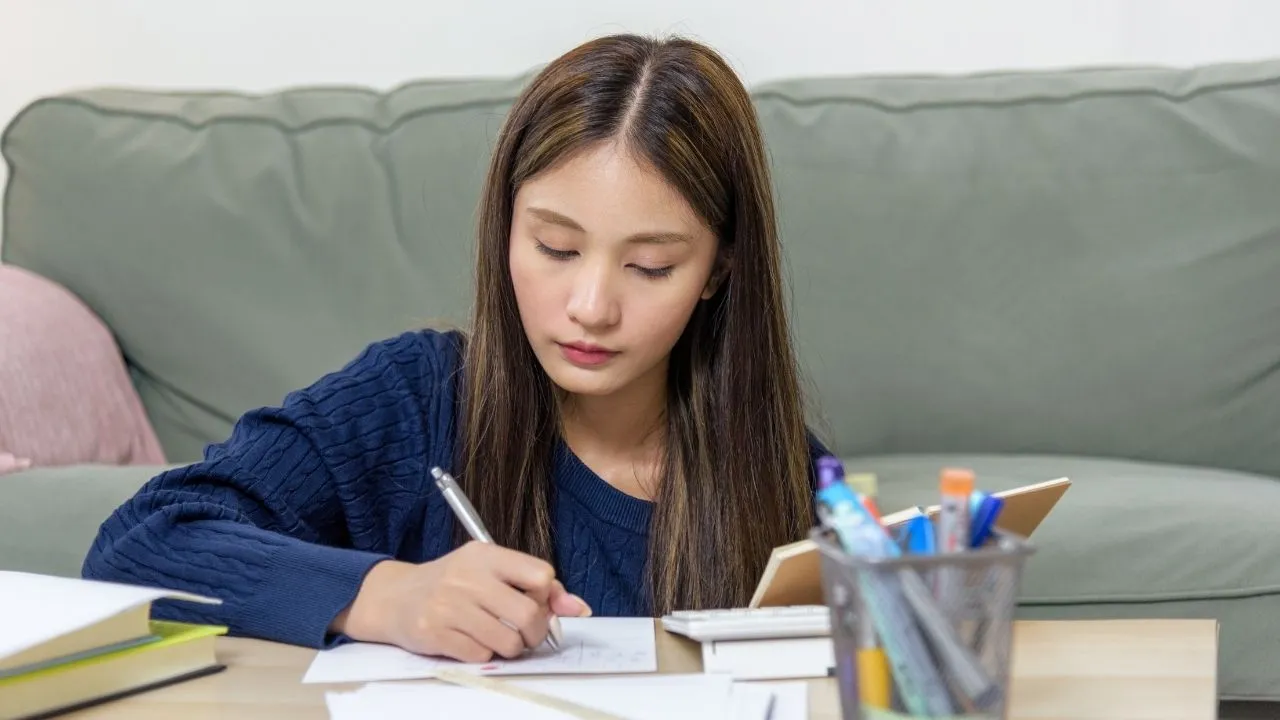 A woman sits at a table, writing on a piece of paper with a focused expression.